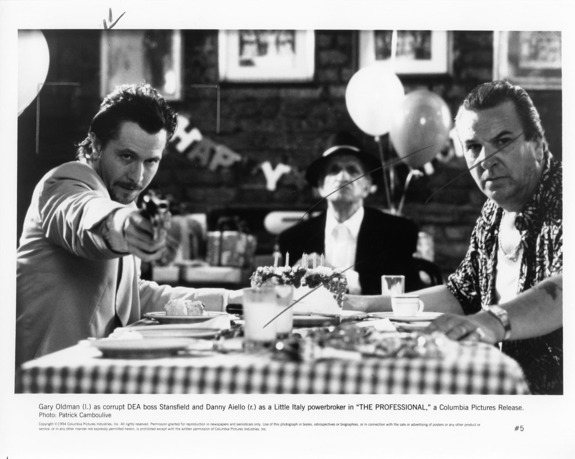Black and white publicity photo from THE PROFESSIONAL featuring Gary Oldman point a gun at us while sitting at a restaurant table with Danny Aiello and another man. A Happy Birthday decoration can be seen in the background.