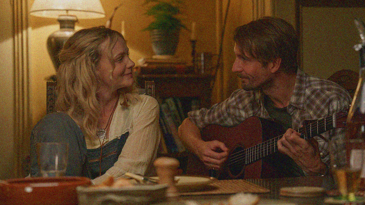 Tom Basden plays guitar at a dining room table while Carey Mulligan sits beside him with a smile, both looking at the other.
