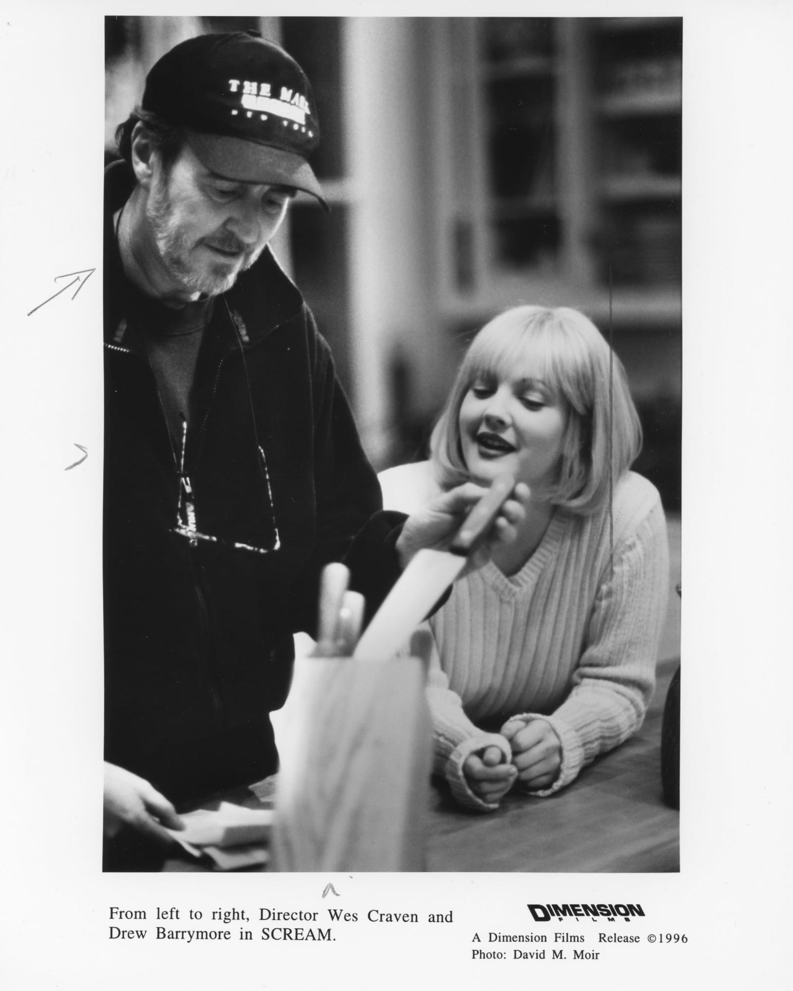 Director Wes Craven (standing at counter with baseball cap, glasses hanging around neck, grabbing knife from block) and Drew Barrymore (leaning on counter next to him) on the set of SCREAM.