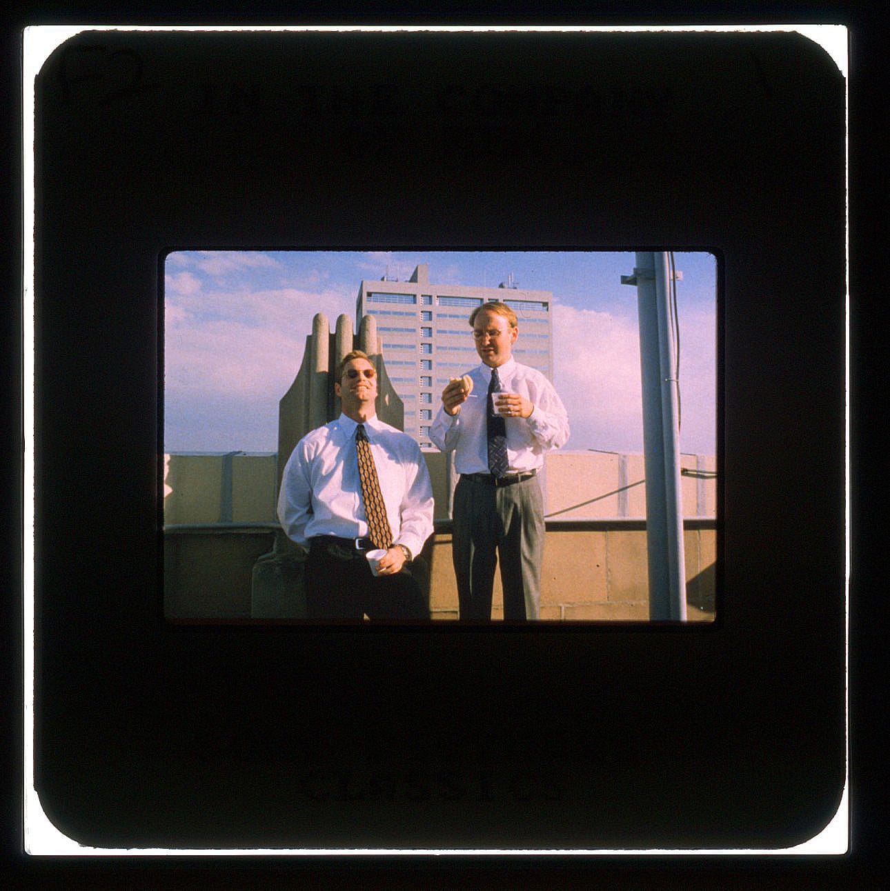 Two men leaning against the ledge of a buildings roof, one grinning with sunglasses and a cup in his hand, the other talking with a bagel in his.