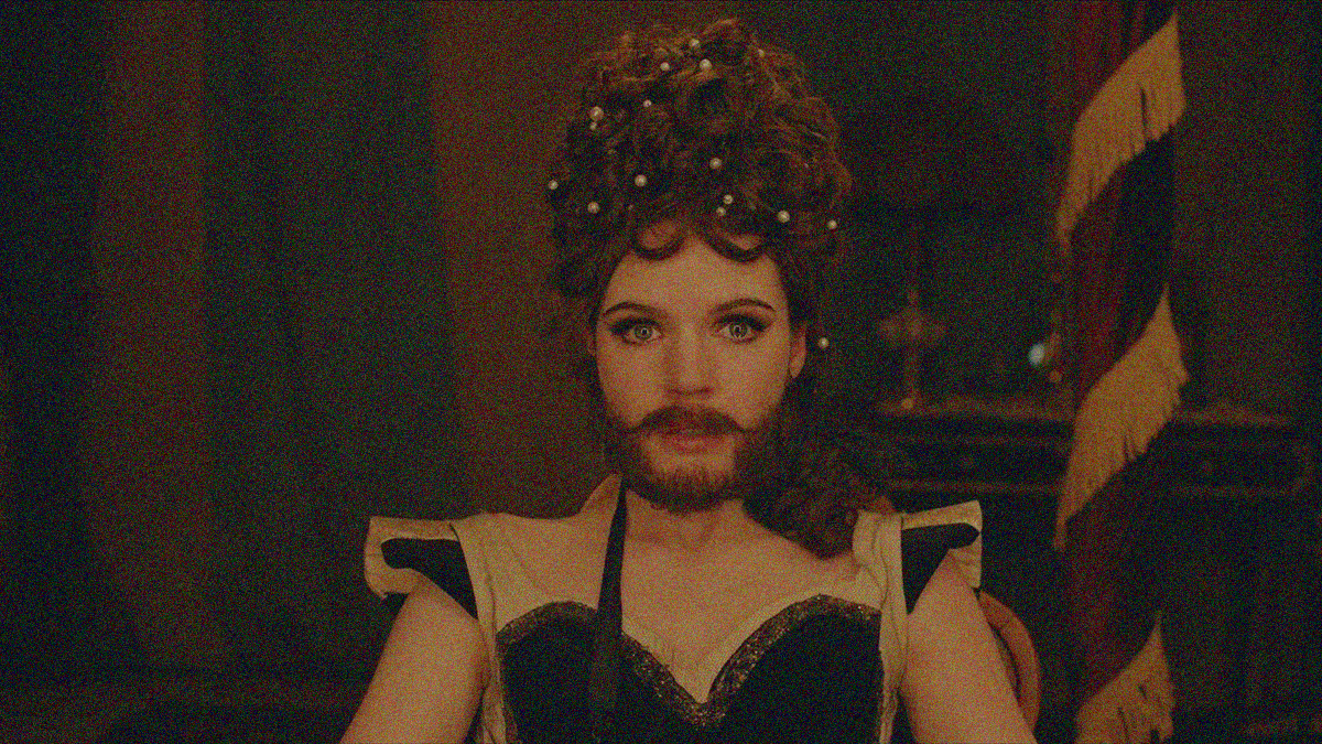A bearded woman with large hair staring at the camera with a wound flag and small table and lamp behind her.