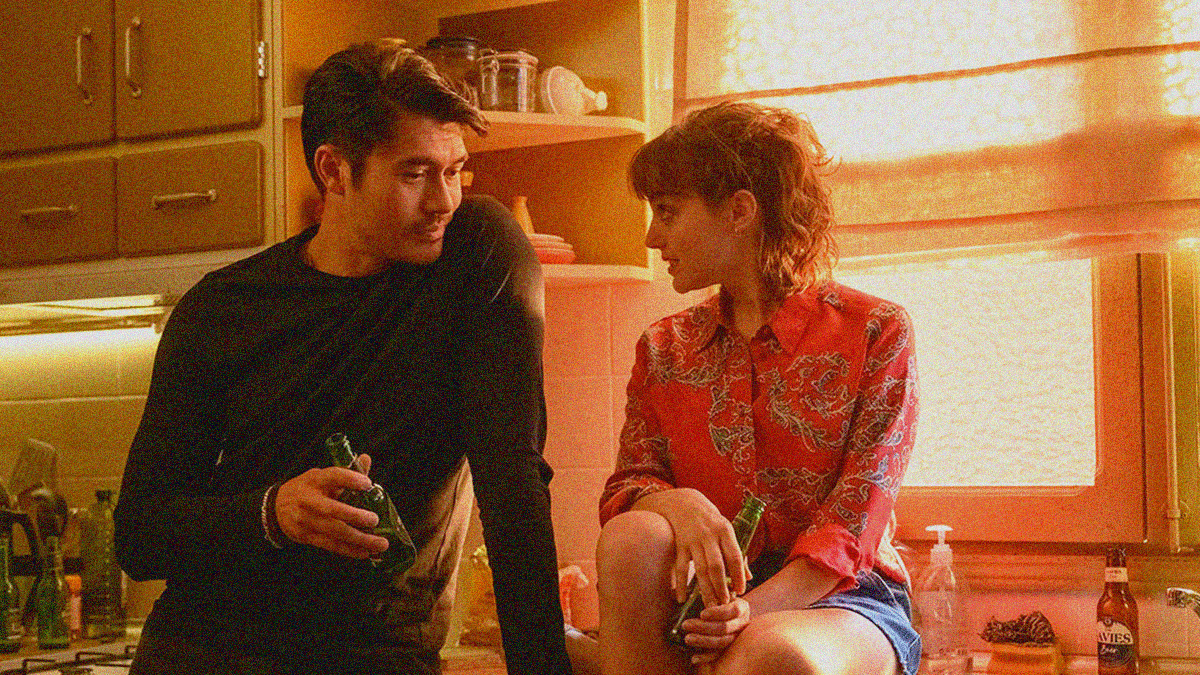 Henry Golding (standing) and Beatrice Grannò (sitting on counter) in a kitchen with beer bottles in hand.