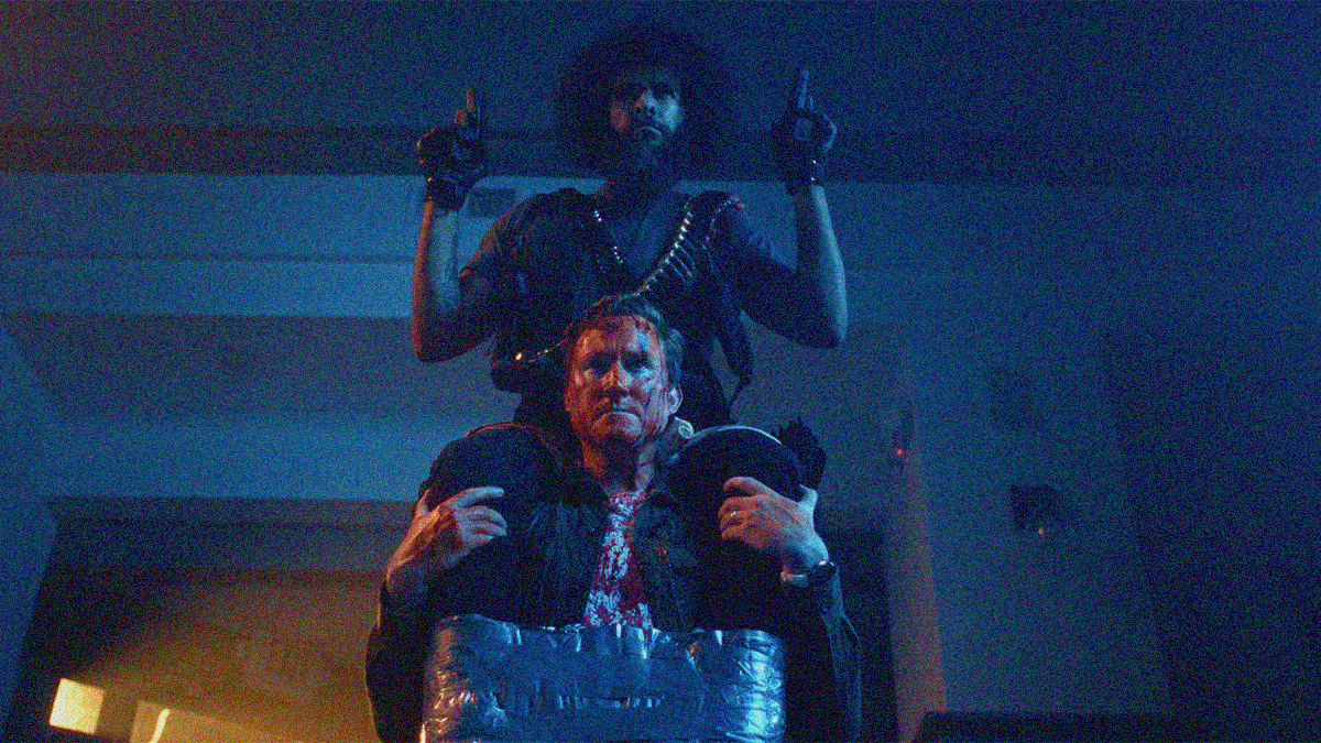 A bearded man with guns in hand and bullets sheathed around his neck is sitting on another man's shoulders with ankles duct taped to his waist.