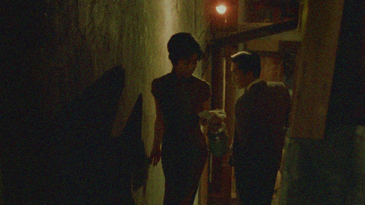 Outdoor alleyway with Maggie Cheung walking towards the screen as Tony Leung walks away from it, both turning back to look at each other.