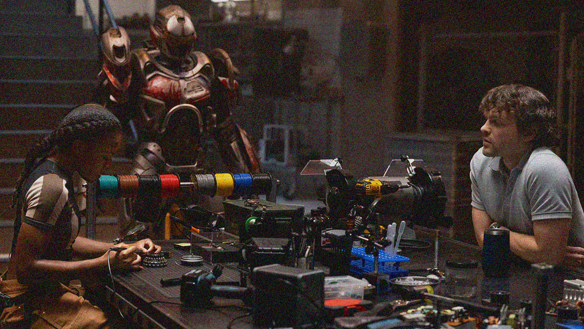 Riri and Joe sit on opposite sides of a table full of tech products and wires while the Ironheart suit stands in the background.