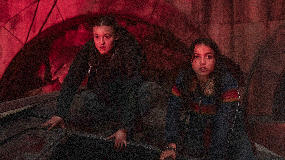 Bella Ramsey and Isabela Merced are crouched on the top of a derelict train car within a darkened tunnel filtered through red light.