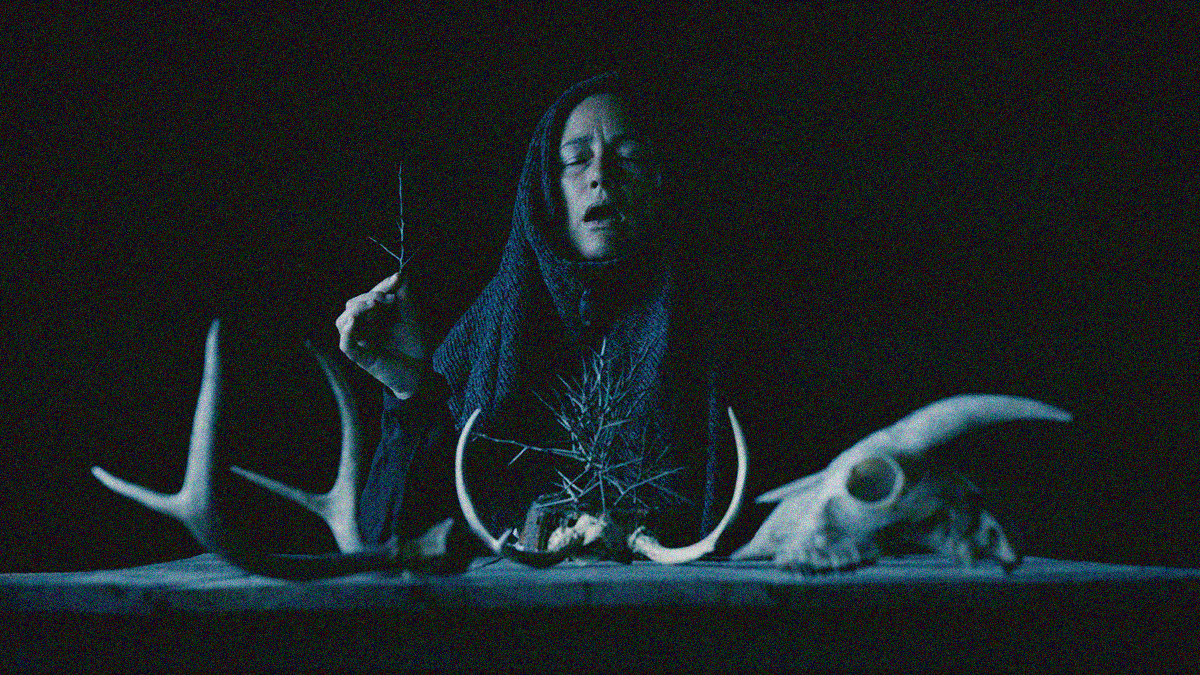 A woman seated in front of a table with antlers and skulls, head covered so her only her face is seen. Her mouth is open and a stick is held between her thumb and index finger.