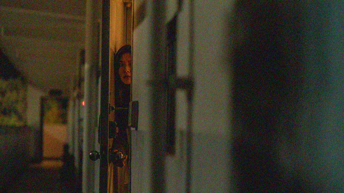 A woman peers out through the crack of her apartment door onto the narrow balcony from an extreme camera angle.