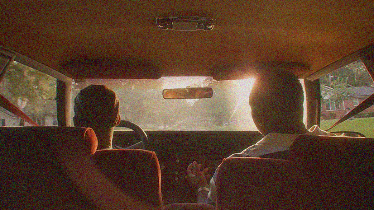 Shot from the backseat of a car with two men conversing in the front seat as sun glare comes through windshield.