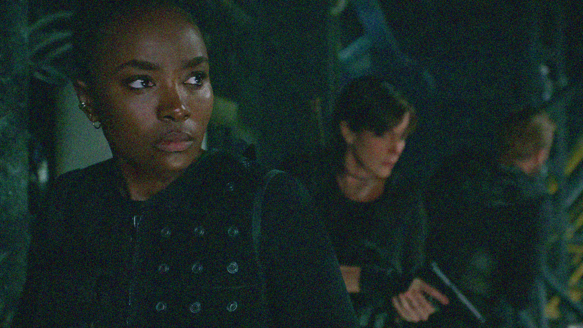 KiKi Layne in the foreground looks off towards right side of the frame while Charlize Theron stands behind her, doing the same with gun drawn.