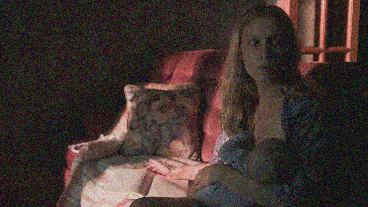 A woman sits on a couch while nursing her baby with a sharp look off-screen.