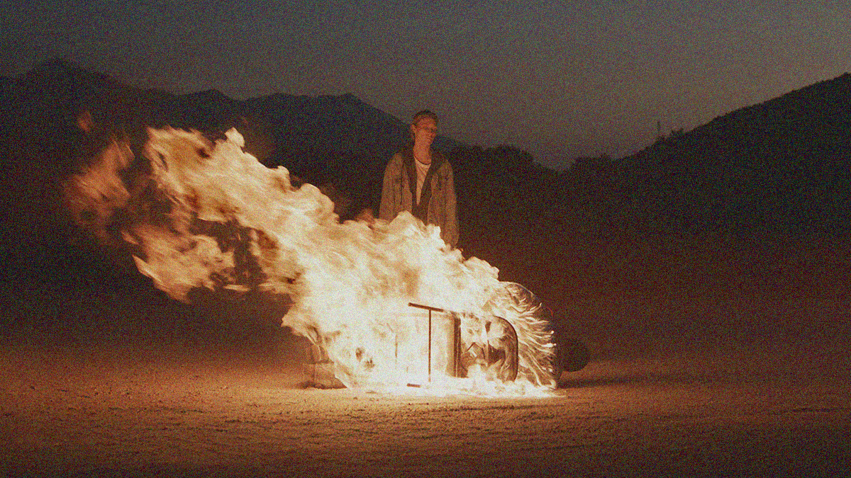 Outdoor, desolate scene at night with mountains in background, a woman stands over a man engulfed in flames while tied to a chair on the ground.