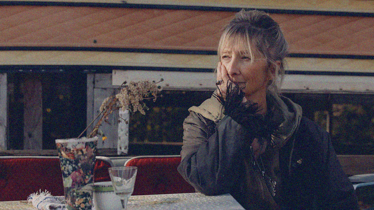 A woman sits outside at a table with head leaning against the palm of her hand.