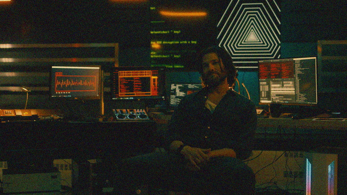 A man sits in front of a large desk with multiple computer screens showing a variety of inputs.
