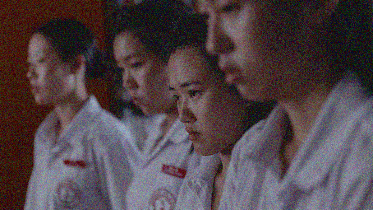 Four teen girls in close up standing shoulder to shoulder looking down as they are reprimanded. Shallow focus keeps the third from the left crisp while the others slightly blur.