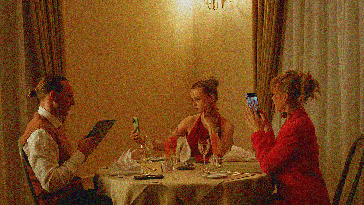 A father looks at the menu while his daughter and wife take a selfie and and scroll their phones respectively at a fancy restaurant.