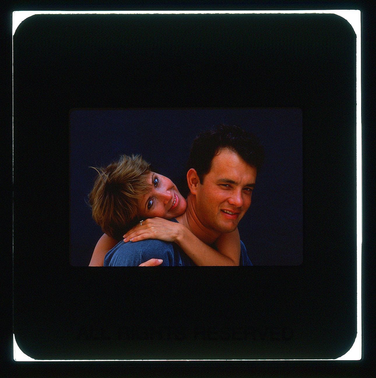 Carrie Fisher and Tom Hanks in an embrace and smiling, her arm wrapped around his neck from behind with head on his shoulder.