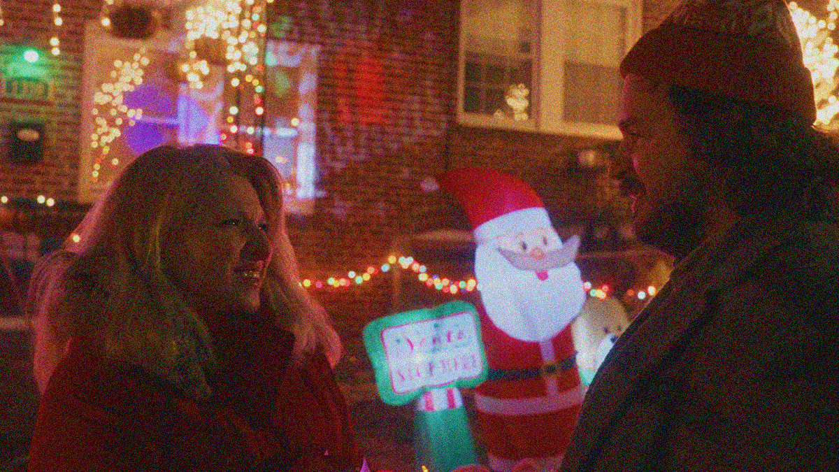 A woman and a man look at each other in winter coast outside of a house decorated for Christmas with a blow-up Santa and North Pole sign.
