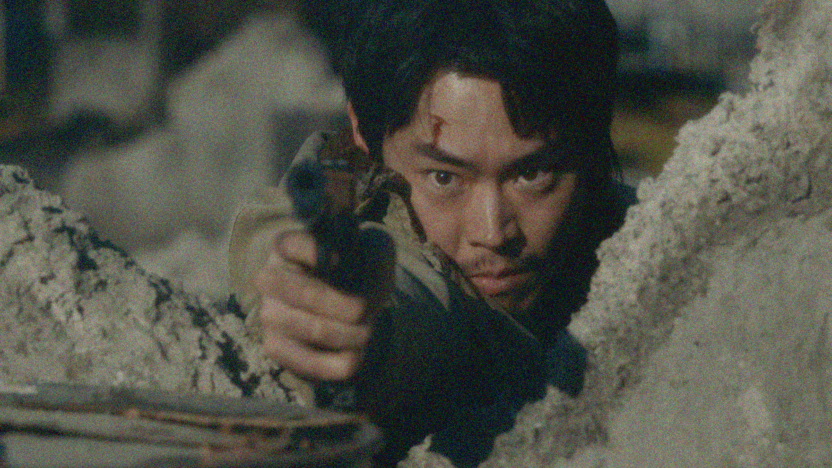 A man is seen between two craggy pieces of concrete pointing a gun towards the camera.