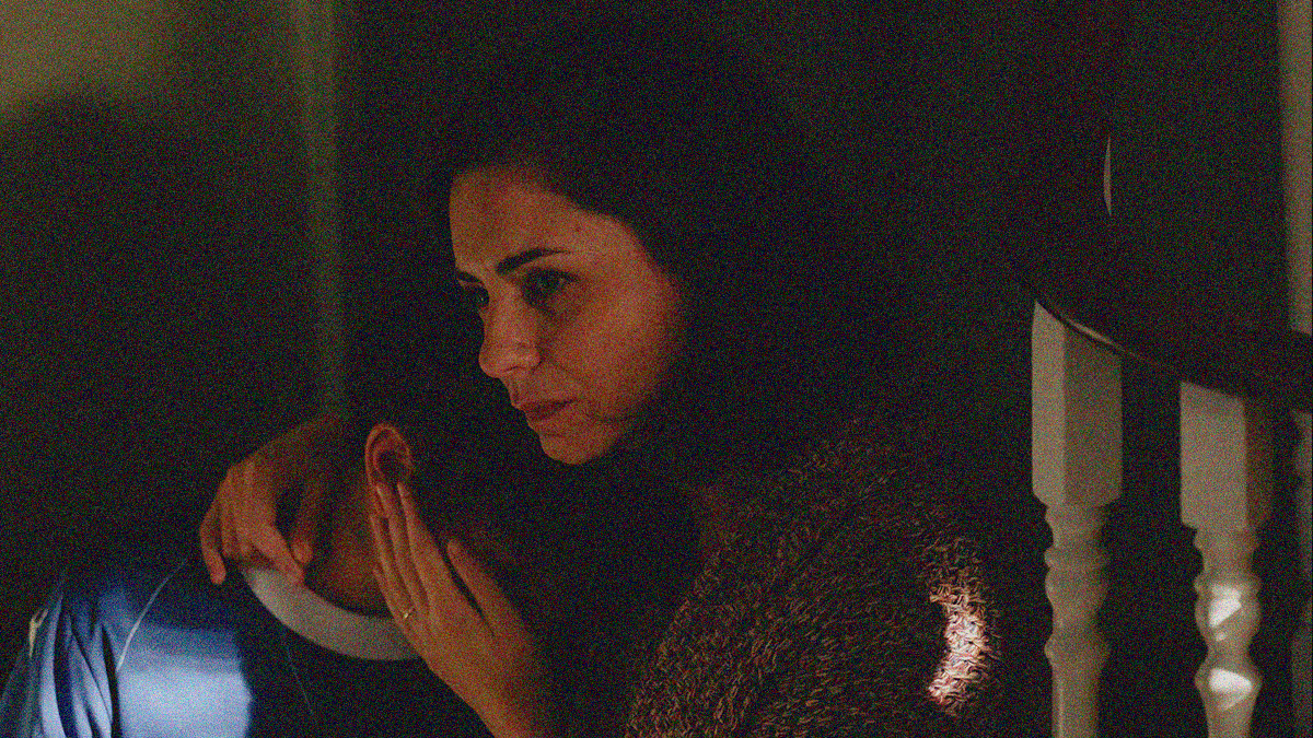 A woman cradles her son's head while sitting on a staircase and looking off-screen with sorrow.