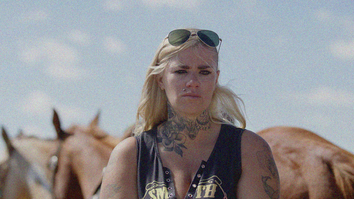 A woman stands in the foreground with a forlorn expression, sunglasses on head, tattoo around neck and on shoulder. Horses are seen in the background.