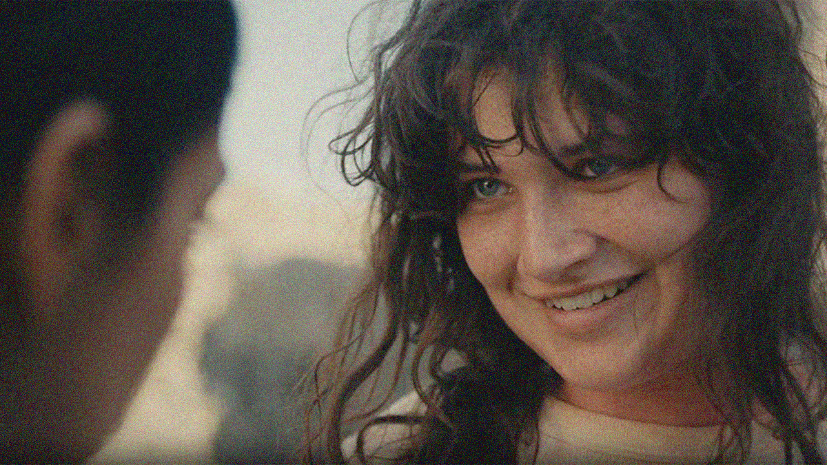 A woman in close-up smiles and fills the right half of the image while looking at a figure whose face barely creeps into frame at left.