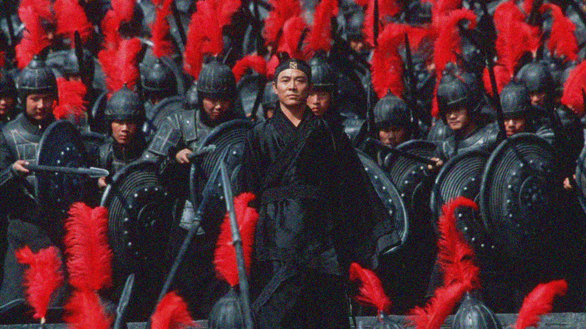 A robed man stoically stands with red-feathered and armored soldiers—spears drawn and shields up—filling the frame above and below him.