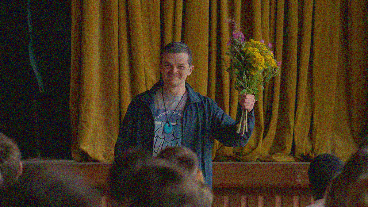 A young man stands in front of a stage with the tops of audience member heads in the foreground. He is smiling and holding up a bouquet of flowers.