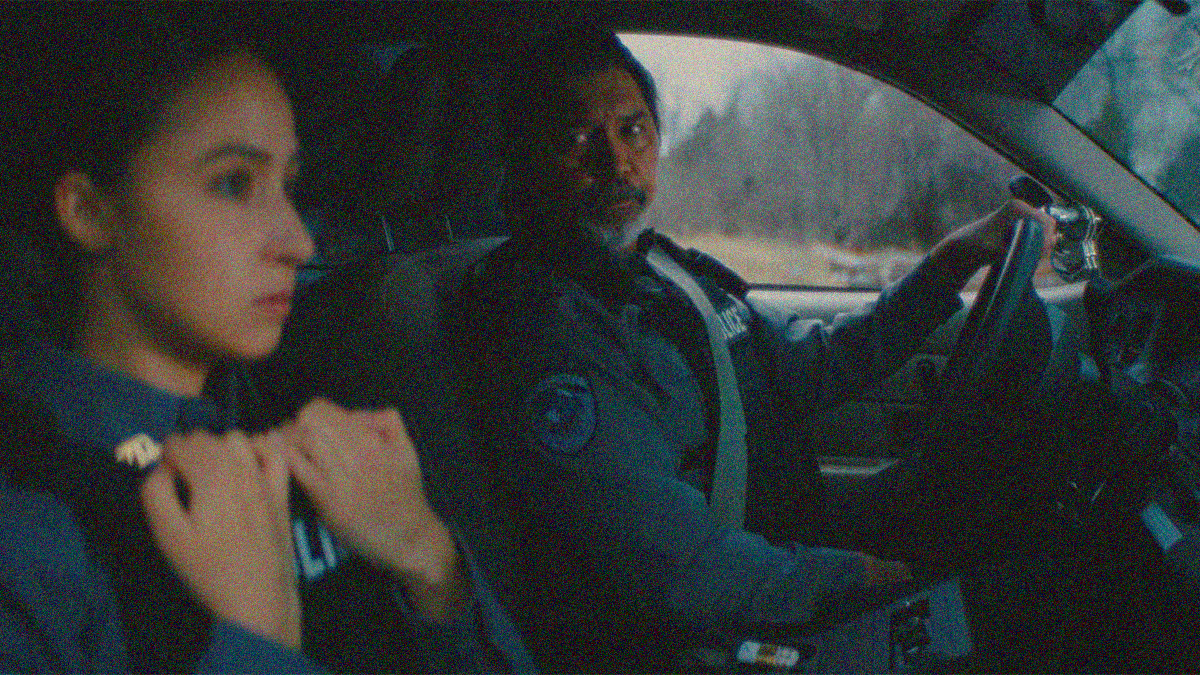 A male cop in the driver's seat of a car in focus in the background looks over at a female cop in the passenger seat not in focus in the foreground.