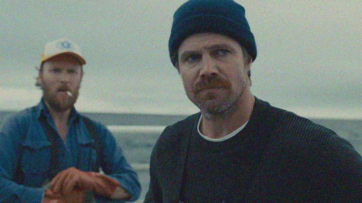 Two men standing on a boat in the ocean in close-up. The one in the background wears a white hat and has a cigarette in his mouth. The one in the foreground wears a black cap and scowls.