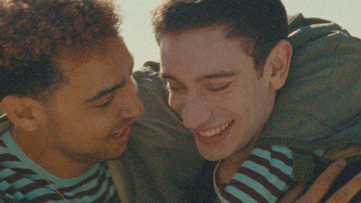 Two men embrace in close-up, the one on left has arm around the shoulder of the one on the right. Both are smiling/laughing.