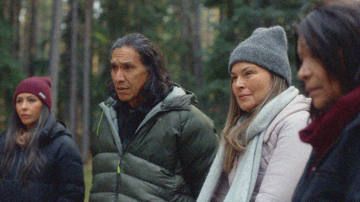 Four adults standing shoulder to shoulder outside with trees in the background. Shallow focus has the man and woman in the center crisp while the women bookending them are blurred.