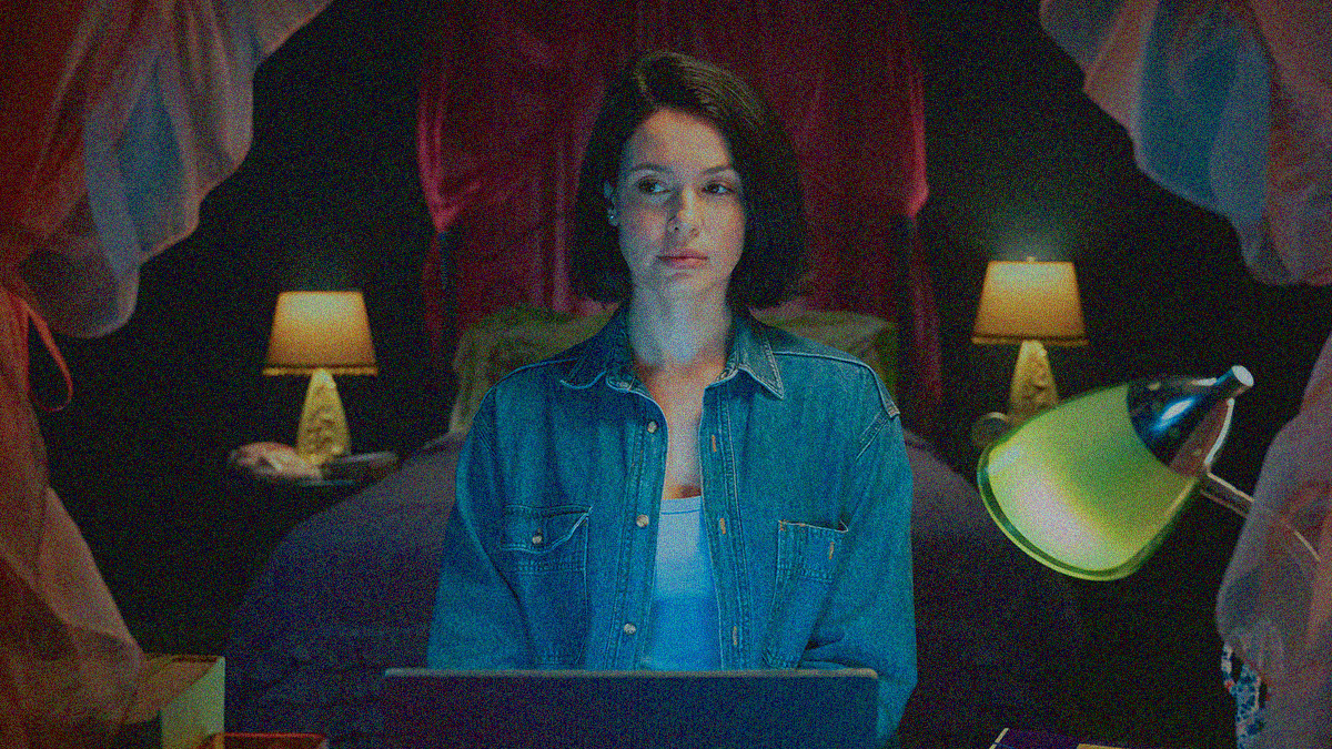 A woman in a demin button down sits in front of a laptop with a bed lit by lamps on either side of it seen behind her.