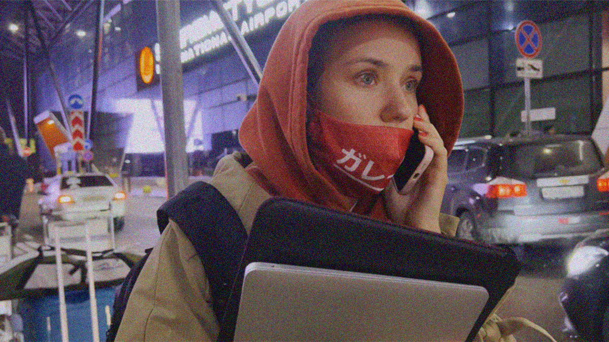 A woman stands outside a busy airport in a red hoodie and red mask (drooped beneath her phone), staring wide-eyed off in the distance while on her cellphone.