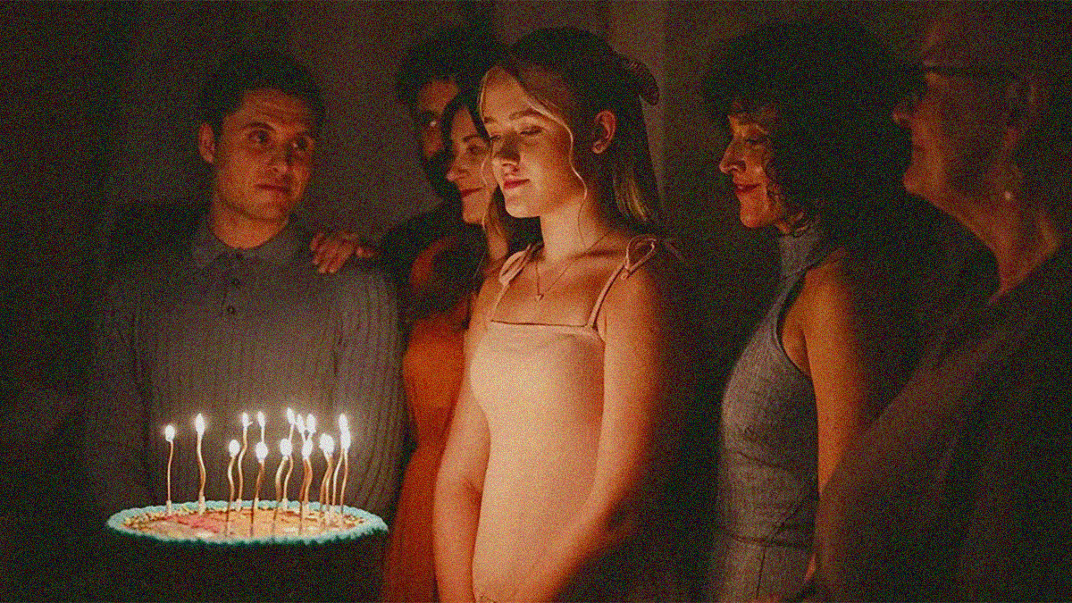 A group of people surround a young girl as she looks down upon a birthday cake with fifteen lit candles.