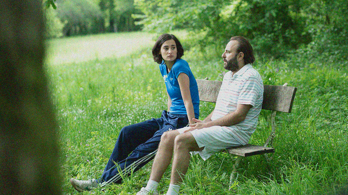 A man and a woman sit on a bench in the country amidst green everywhere (grass, trees, etc.)