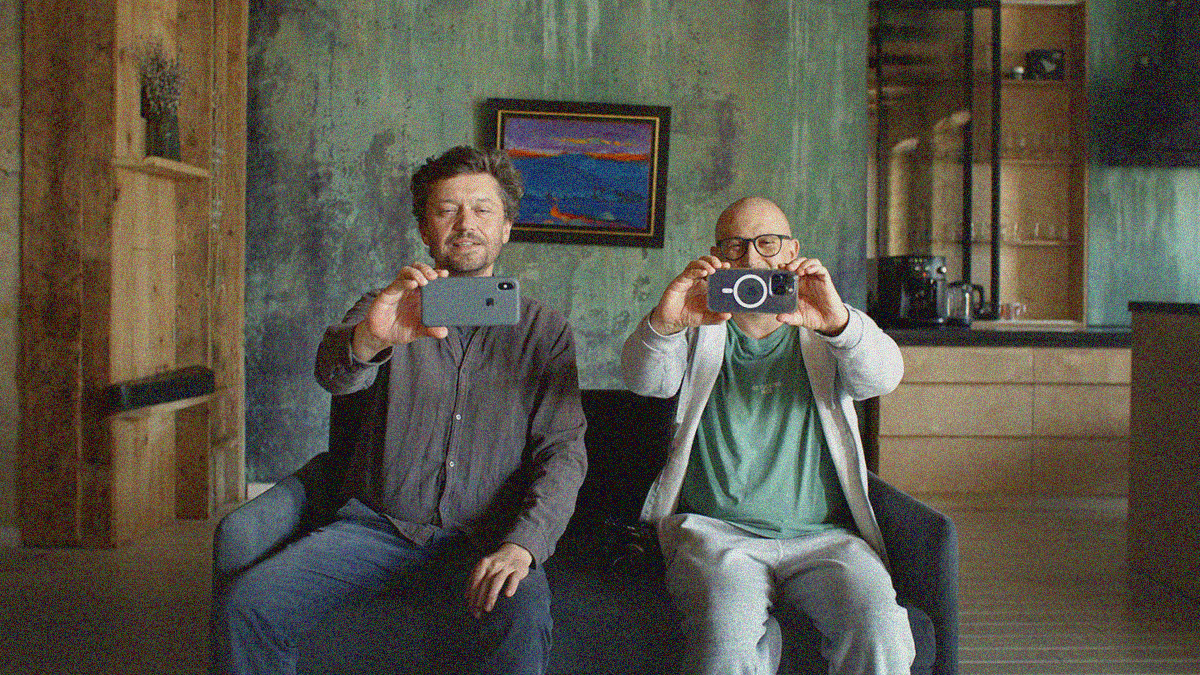 Two men sitting on a couch with a concrete wall adorned by a painting of water behind them are smiling and staring into their phones—outstretched and landscape to film themselves.