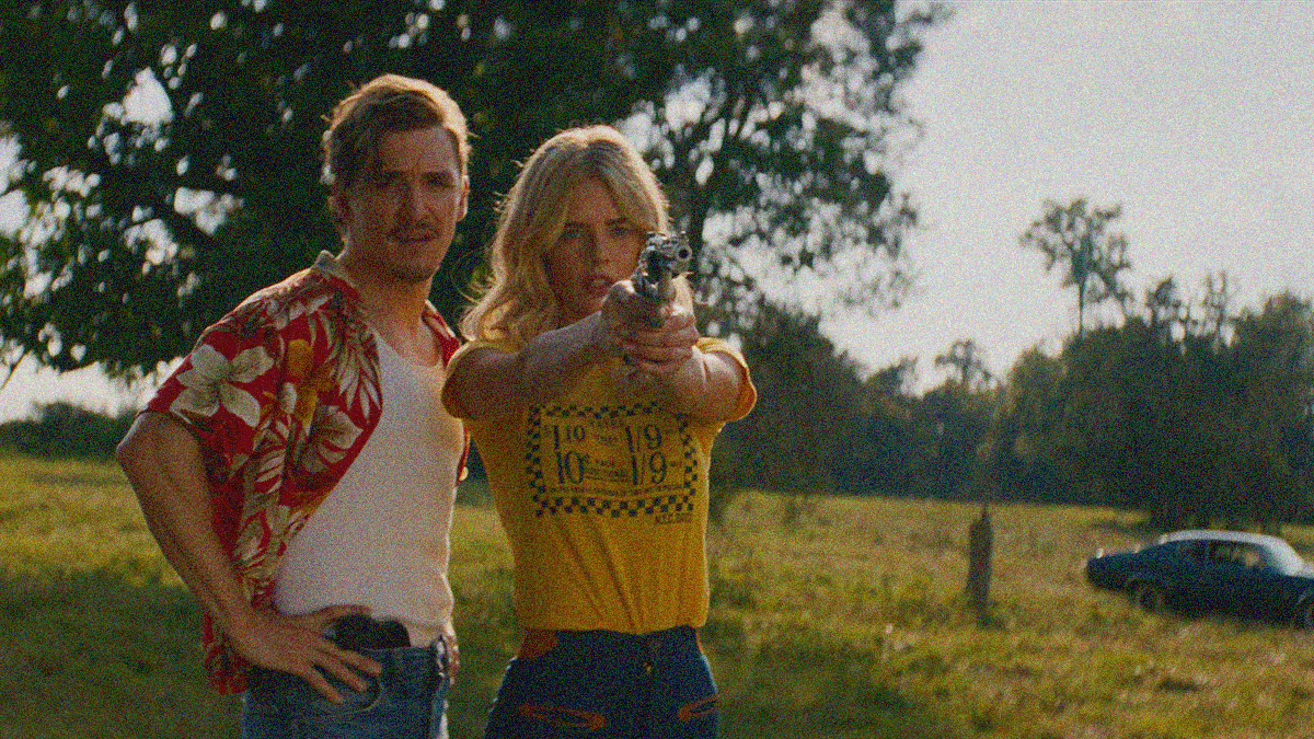A man stands behind a woman as she readies to shoot the gun she has in her outstretched arms.