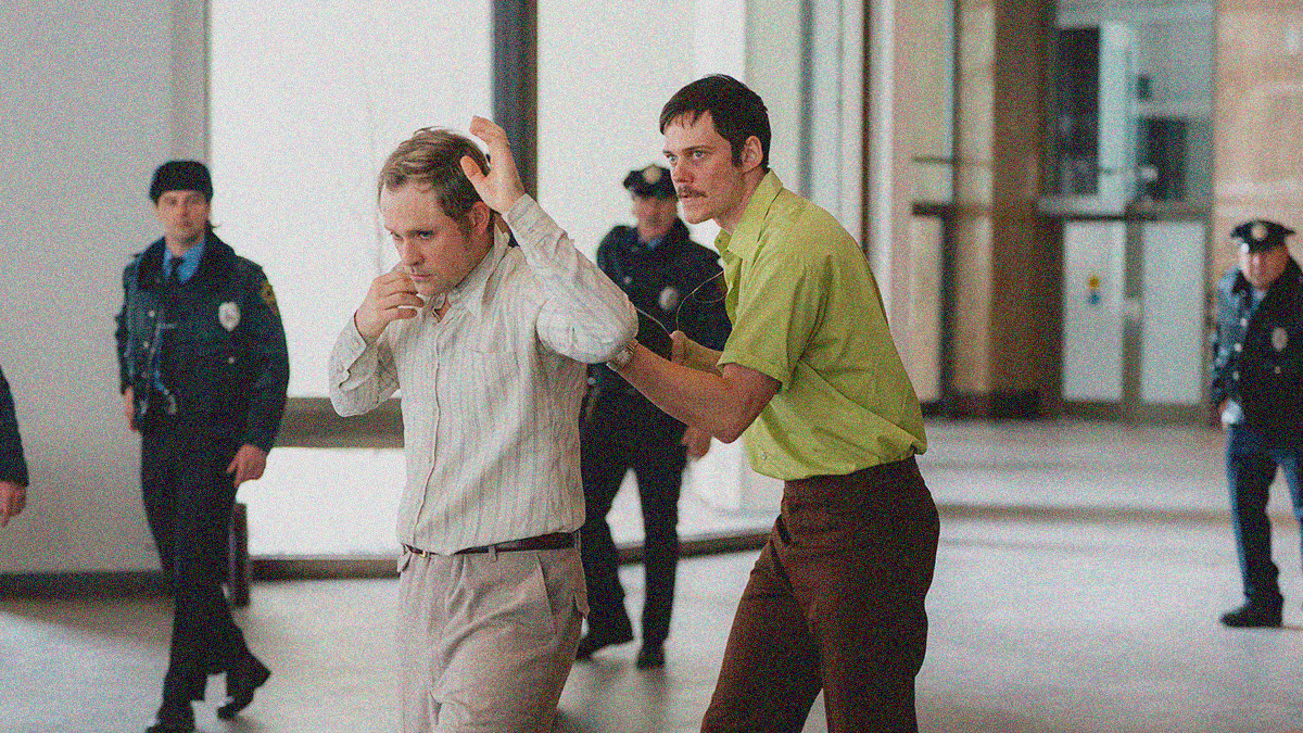 A man in a yellow collared shirt leads another out of a building with a shotgun held to his back. Police officers watch from behind them.