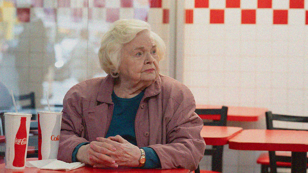 An older woman sits at a fast food restaurant table with hands folded, looking off to the right.