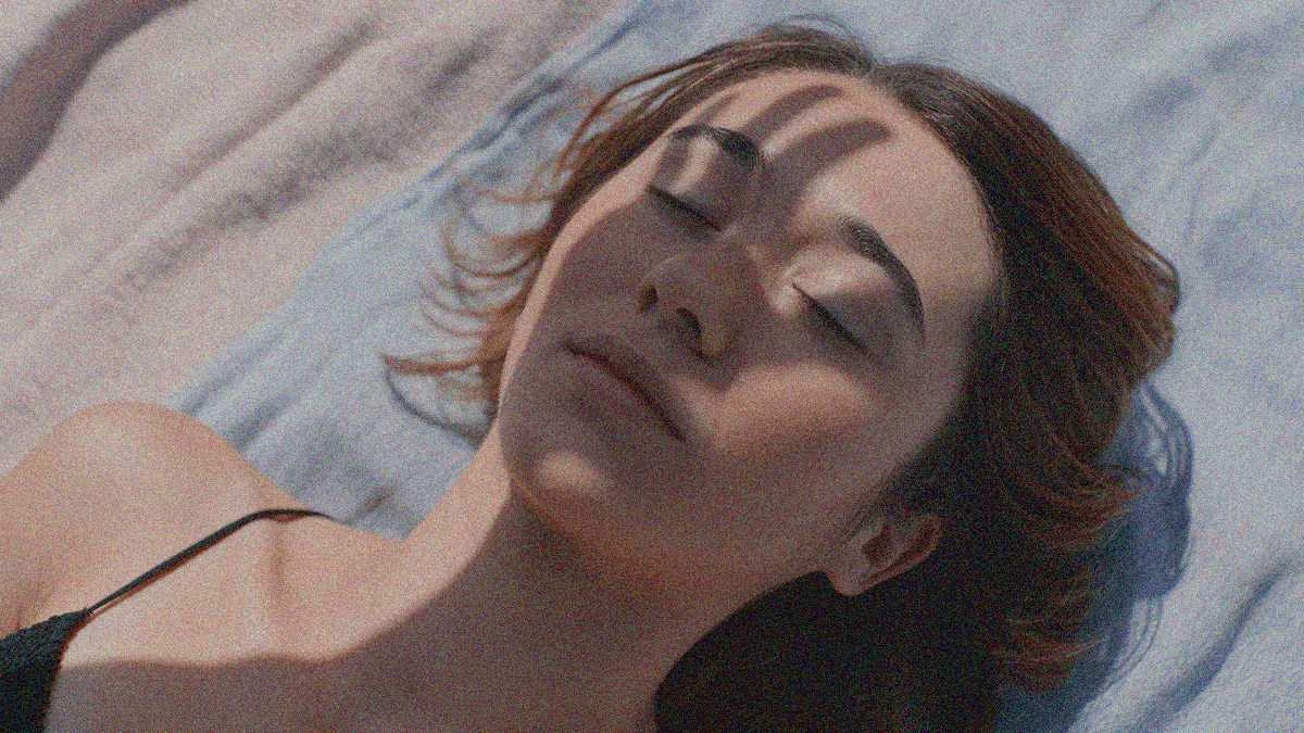 A close-up of a young woman lying on a beach towel with the shadow of someone else's hand covering her face.