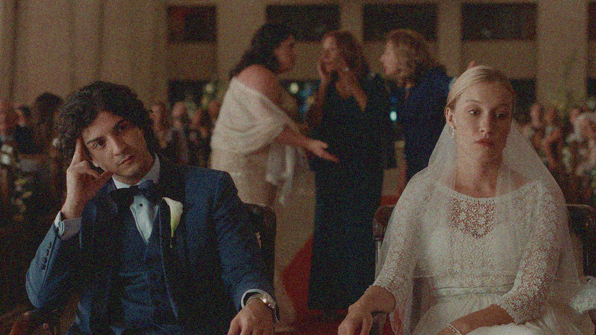 A groom and bride sit in the foreground facing us with their backs to a full church—looking frustrated and angry as three women stand and argue behind them in the aisle.