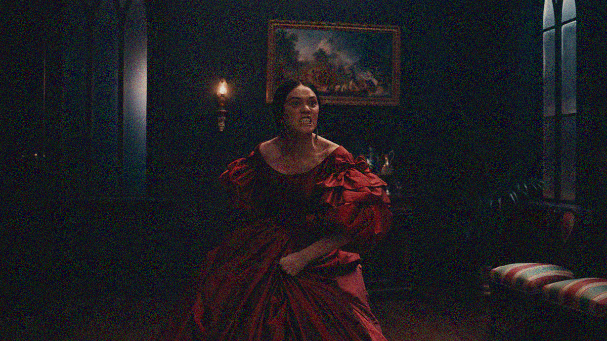 A Māori woman in a red puffy Victorian dress performs a haka in the dimly lit room of a mansion.