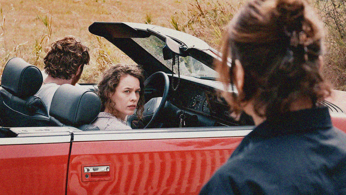 A woman in the mid-ground is turned towards us while sitting in a convertible. She's staring at someone in the foreground for whom we only see the back of the head.