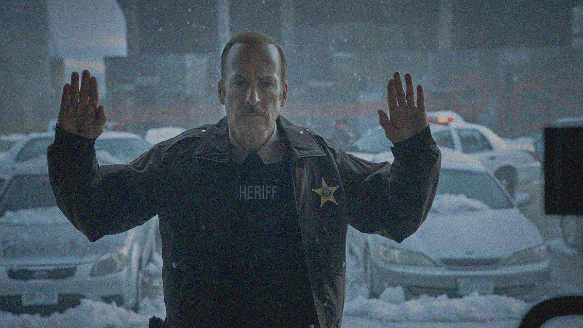 A sheriff stands outside with arms raised in surrender. Snow covered cars are seen behind him.