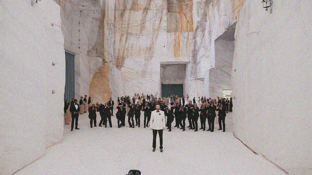 A long shot between two giant slabs of rock with a man in a white tux facing us as a collection of black suited photographers line-up behind him.