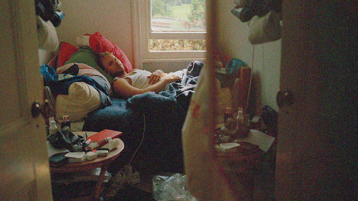 Peering through an open door, we see a young man lying in bed looking at the camera, his room is a mess.