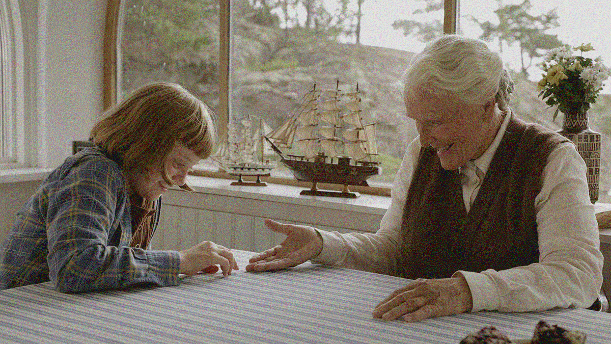 A young girl and an older woman sitting and laughing at a table with hands outstretched. Model ships ate on a windowsill behind them.