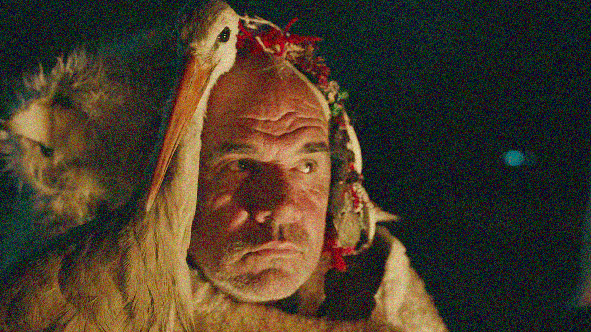 Close-up of an older man looking off-screen with a stork standing and staring at the camera from just in front of his face.