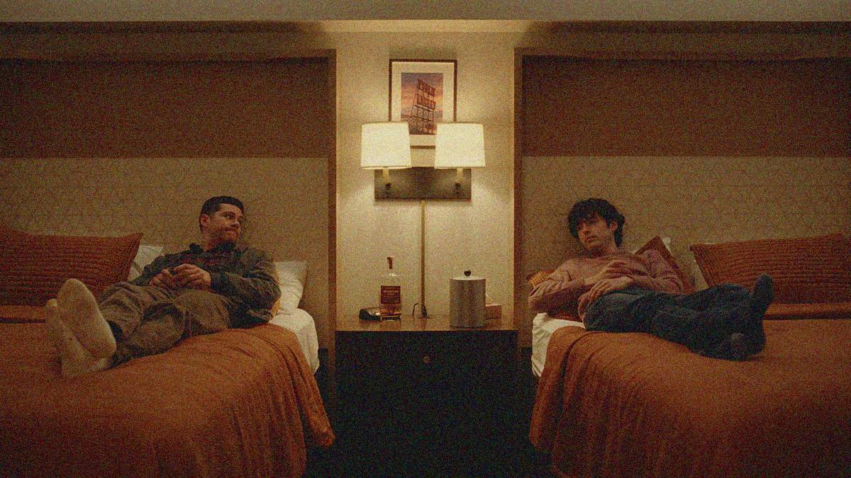 Two men laying on the covers of separate beds in a hotel room looking towards the other. Wall lamps are lit between them.